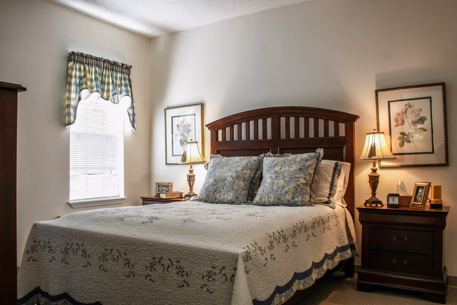 A neatly made bedroom with a wooden headboard, floral pillows, bedside tables with lamps, framed art, and a window with a valance.