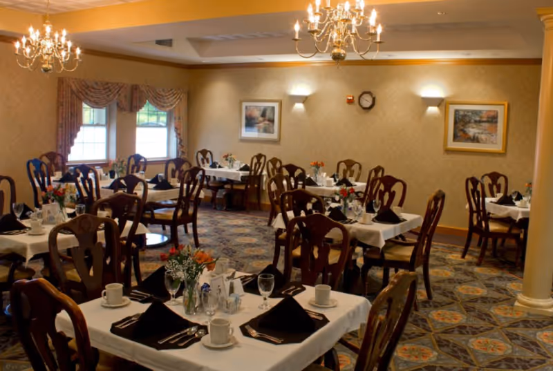 A dining room with multiple tables set for a meal, each table covered with a white tablecloth, black napkins, cups, glasses, and silverware. The room features wooden chairs, chandeliers hanging from the ceiling, patterned carpet, framed paintings on the walls, and windows with decorative curtains.