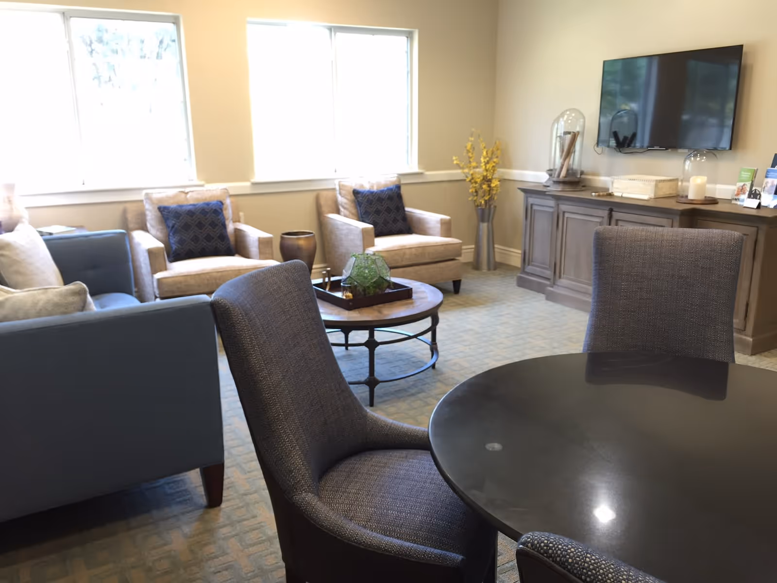 A cozy living room area with two beige armchairs with dark blue patterned pillows, a blue sofa, a round black table with two gray upholstered chairs, a round wooden coffee table with a decorative tray, a wooden cabinet with decorative items, and a wall-mounted flat-screen TV. Two large windows let in natural light.
