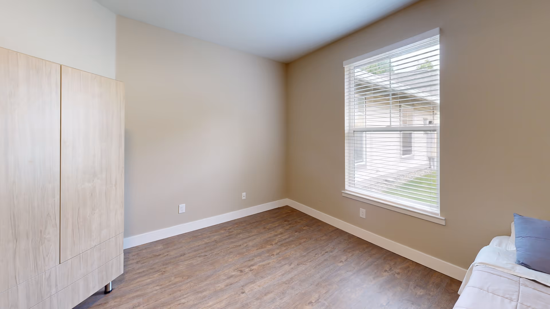 A small, simple bedroom with beige walls, wood flooring, a window with white blinds, a light wood wardrobe on the left, and a bed with white bedding and a blue pillow on the right.