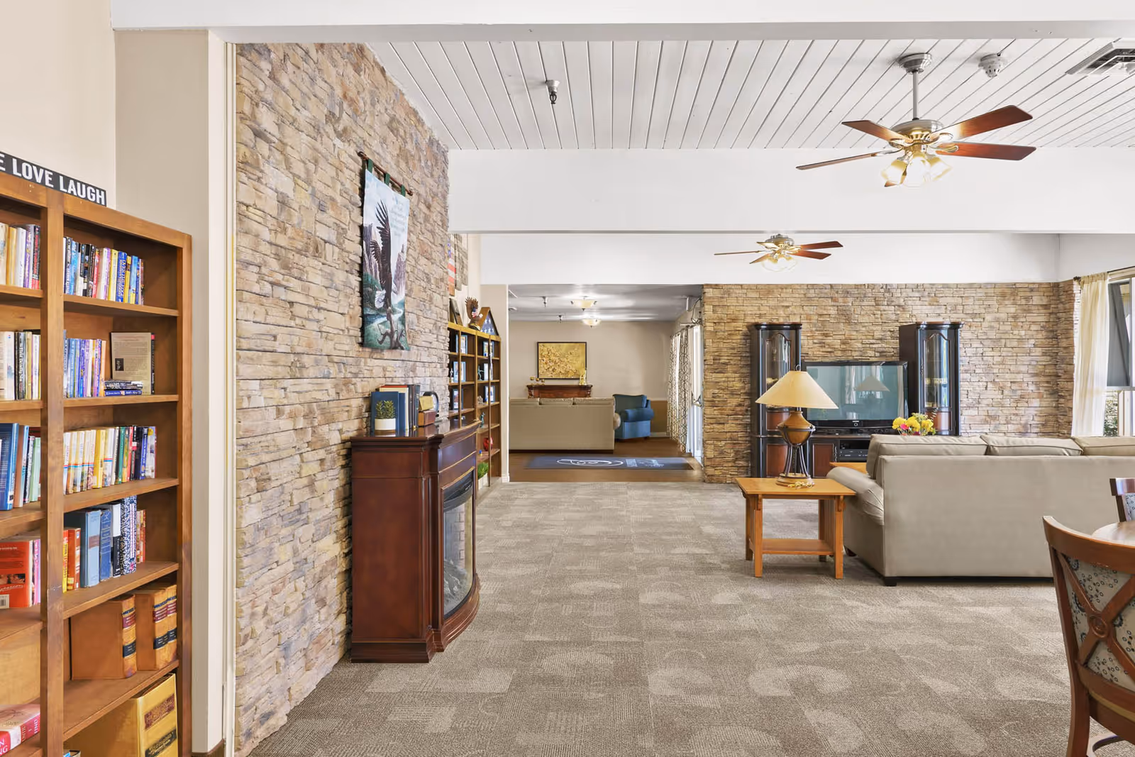 Spacious senior living common room with bookshelves, stone-accent walls, sofas, ceiling fans, and a TV area.