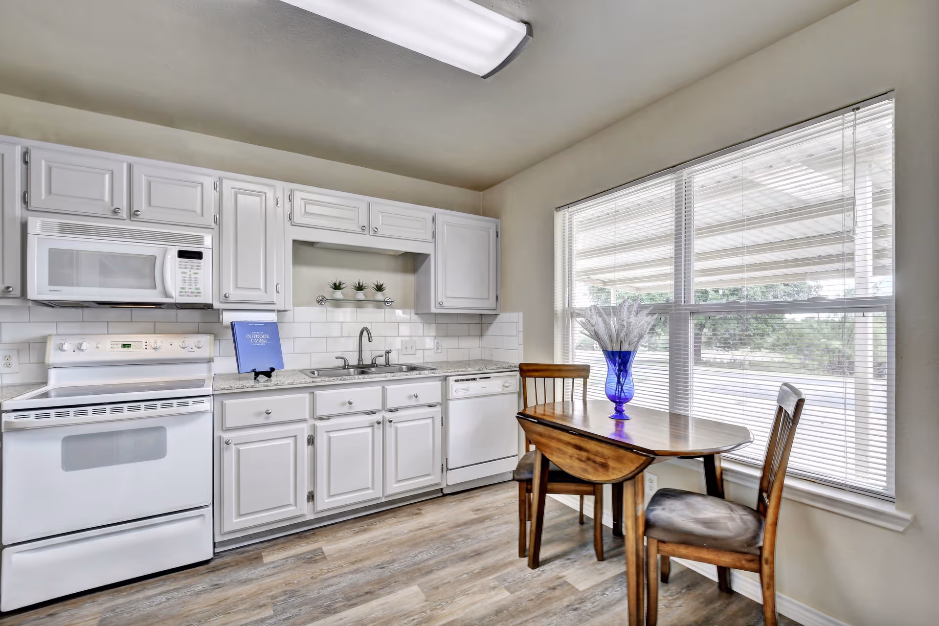 A bright kitchen with white cabinets, a white stove, microwave, and dishwasher. There is a small wooden dining table with two chairs next to a large window with blinds. A blue vase with decorative white branches is placed on the table.