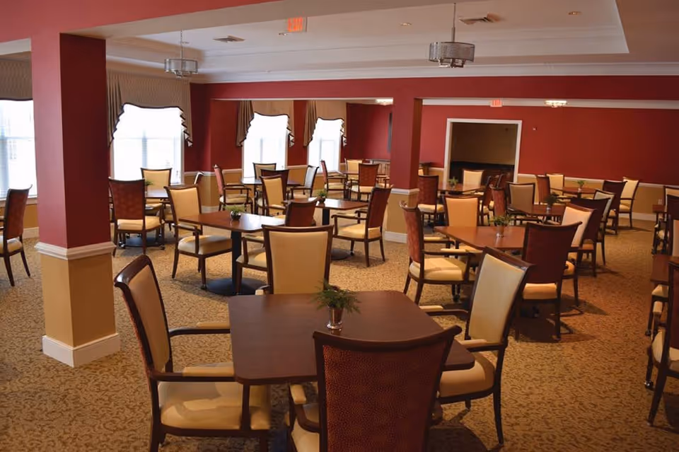 A spacious dining room with multiple square wooden tables and cushioned chairs arranged neatly. The room has red walls with beige wainscoting, large windows with valances allowing natural light, and carpeted flooring. Small potted plants are placed on each table.