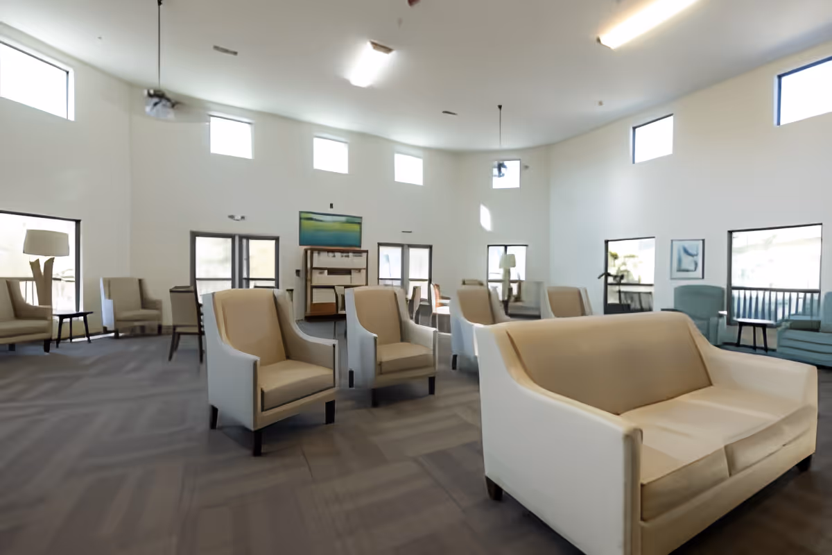 A spacious, well-lit common area with multiple beige armchairs and sofas arranged on a carpeted floor. The room has high ceilings with several small windows near the ceiling and larger windows along the walls, allowing natural light to enter. There are a few pieces of artwork on the walls and a wooden shelving unit in the background.