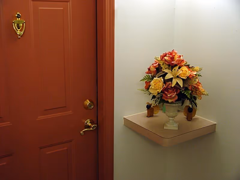 A corner of an interior space featuring a closed reddish-brown door with a gold-colored door knocker and handle. Adjacent to the door is a small wall-mounted shelf holding a decorative vase filled with an arrangement of artificial flowers in shades of red, yellow, and orange.