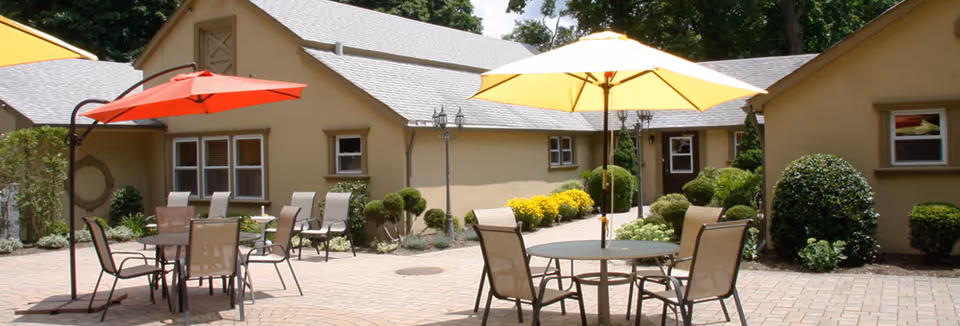 Outdoor patio area at Maryville Enhanced Assisted Living Center with round tables and chairs under large red and yellow umbrellas, surrounded by trimmed bushes and a beige building in the background.