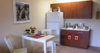 A small kitchen area with a white refrigerator, wooden cabinets, a countertop with a coffee maker and other kitchen items. A white dining table with two chairs is set with placemats and a small flower arrangement. A framed picture hangs on the wall above the table.