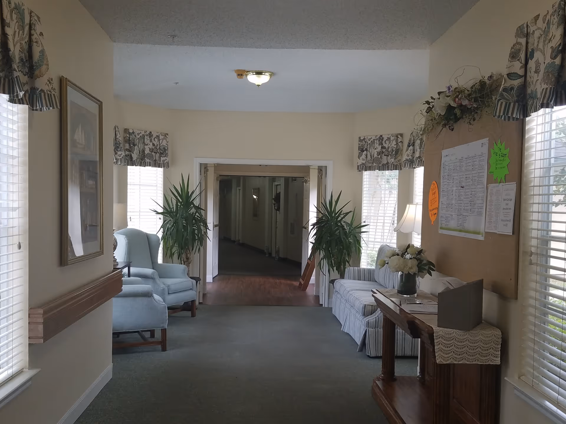 A quiet hallway in a senior living facility with two light blue armchairs on the left, a striped sofa on the right, two potted plants, a wooden table with a lace runner and a vase of flowers, and a bulletin board with notices on the right wall. The hallway leads to a corridor with closed doors.