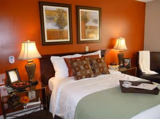 A cozy bedroom with a neatly made bed featuring white sheets, patterned pillows, and a green blanket. Two bedside tables each hold a lamp and decorative items. The wall behind the bed is painted orange and adorned with two framed pictures of trees.