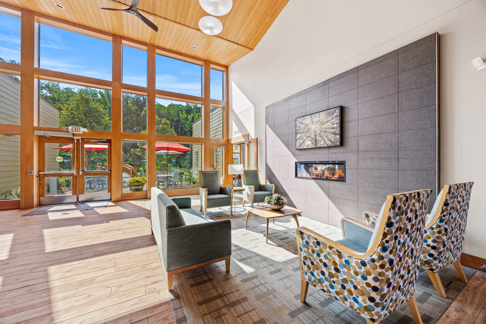 A bright and spacious living room area with large floor-to-ceiling windows letting in natural light. The room features modern furniture including two patterned armchairs, two green armchairs, and a green sofa arranged around a wooden coffee table. A contemporary fireplace is built into a large gray tiled wall with a piece of artwork above it. Outside the windows, there is a patio with red umbrellas and greenery.