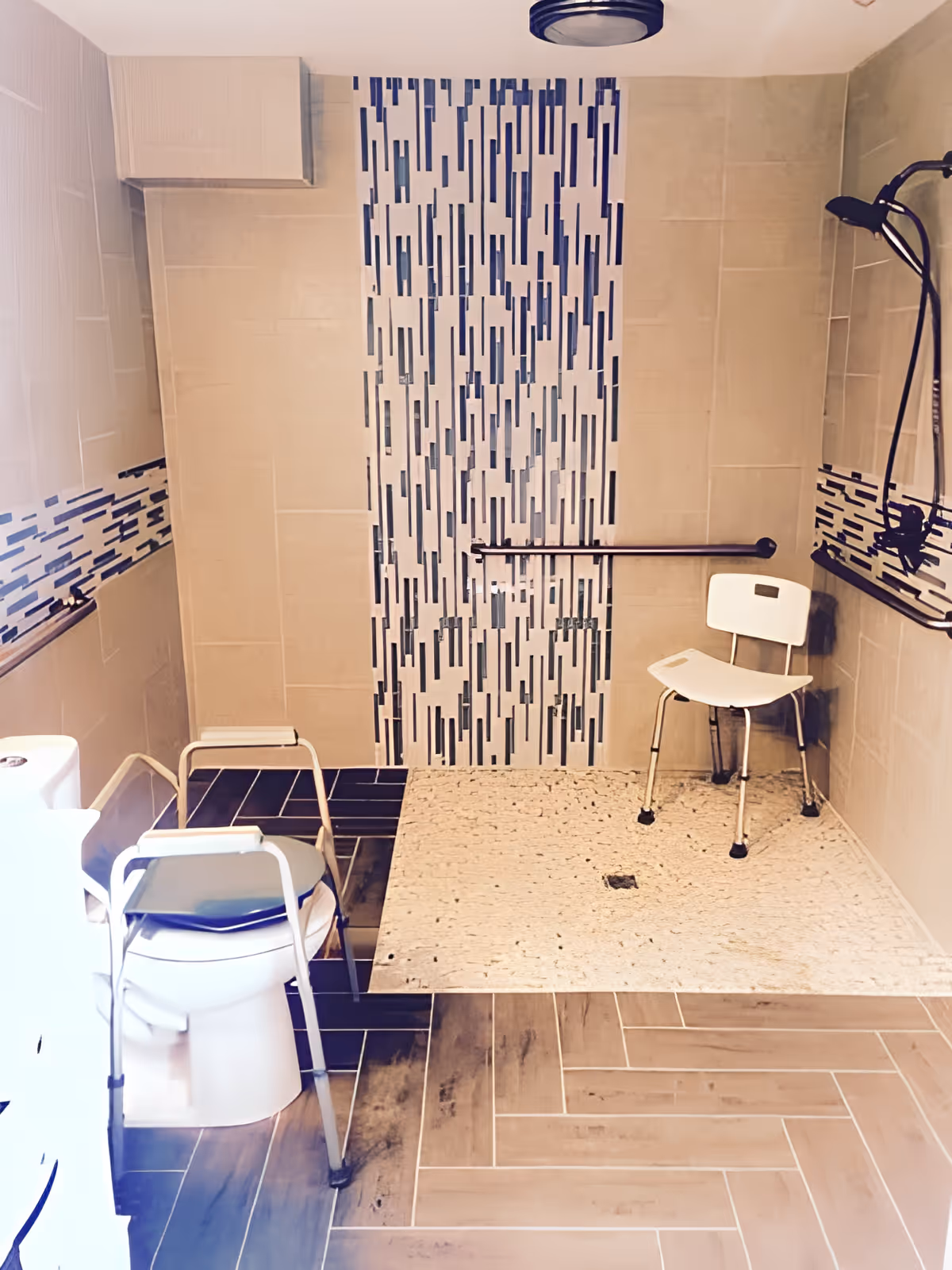 Accessible bathroom with a walk-in shower featuring a white shower chair and a black handheld showerhead. The shower area has a textured floor and vertical blue and white tile accents on the wall. There is a white toilet with a metal walker placed in front of it. The bathroom walls are tiled in beige with a horizontal strip of blue and white tiles. The floor is covered with wood-look tiles arranged in a herringbone pattern.