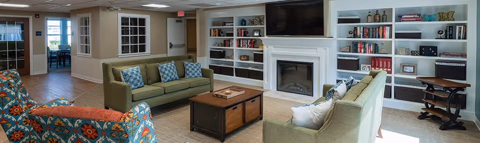 A cozy living room area with two green sofas facing each other, a wooden coffee table in the center, and a colorful patterned armchair. The room features built-in white bookshelves filled with books and decorative items, a fireplace with a mounted flat-screen TV above it, and large windows letting in natural light.