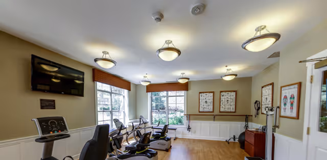 A well-lit exercise room with several stationary recumbent bikes facing large windows that let in natural light. The walls are beige with white wainscoting, and there are framed posters and exercise equipment along the walls. Ceiling lights provide additional illumination.