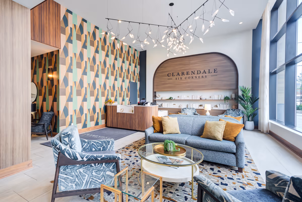 A modern and stylish lobby area of Clarendale Six Corners featuring a gray sofa with mustard and orange pillows, patterned armchairs, a glass coffee table, a colorful geometric patterned wall, a wooden reception desk, and large windows allowing natural light to fill the space.