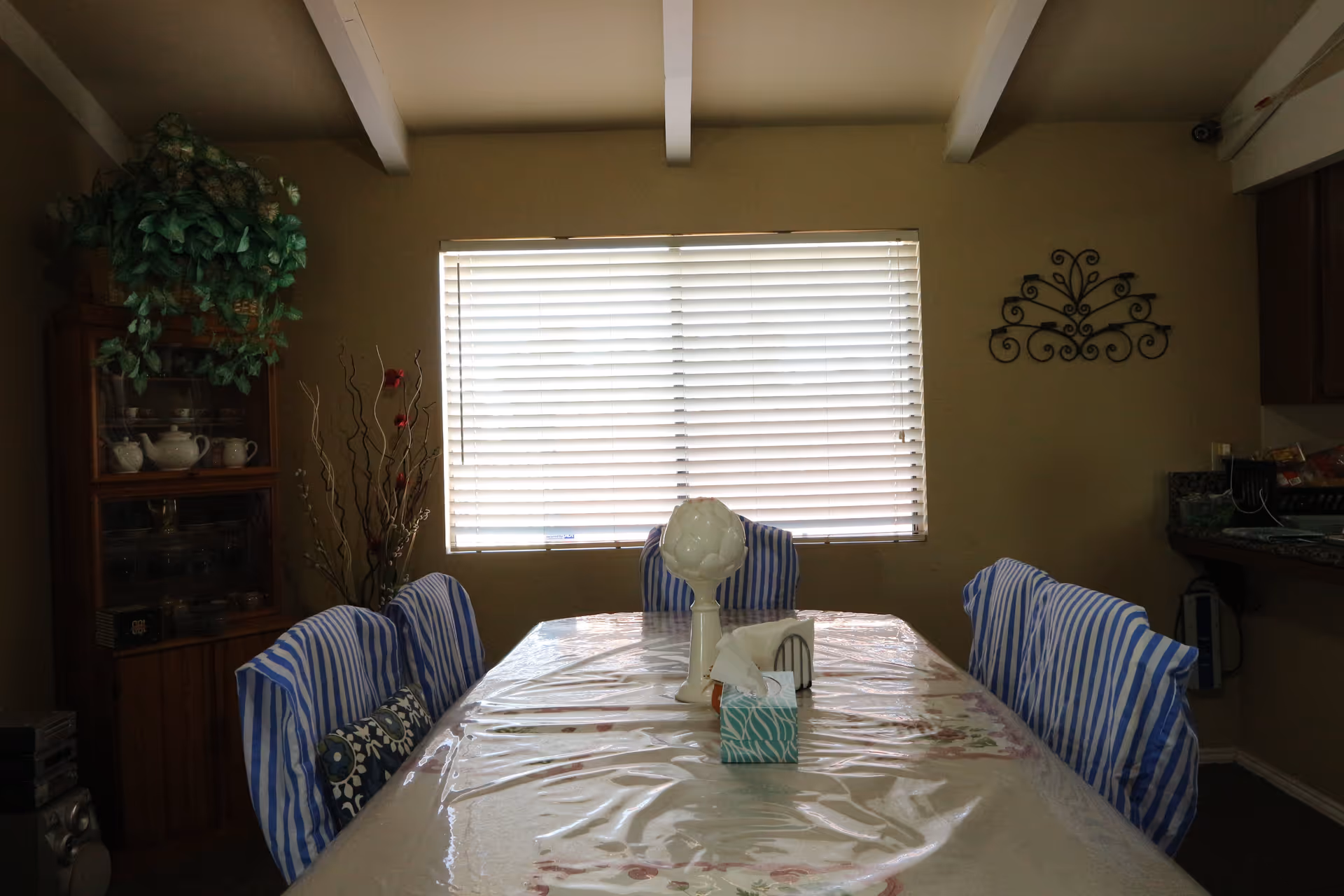 A dining room with a rectangular table covered with a floral tablecloth and a clear plastic cover. Six chairs with blue and white striped covers surround the table. On the table, there is a white decorative object, a tissue box, and a napkin holder. A window with closed blinds is on the wall behind the table. To the left, there is a wooden cabinet with glass doors displaying teapots and cups, topped with a green leafy plant. On the right wall, there is a decorative metal wall hanging and part of a kitchen counter is visible.