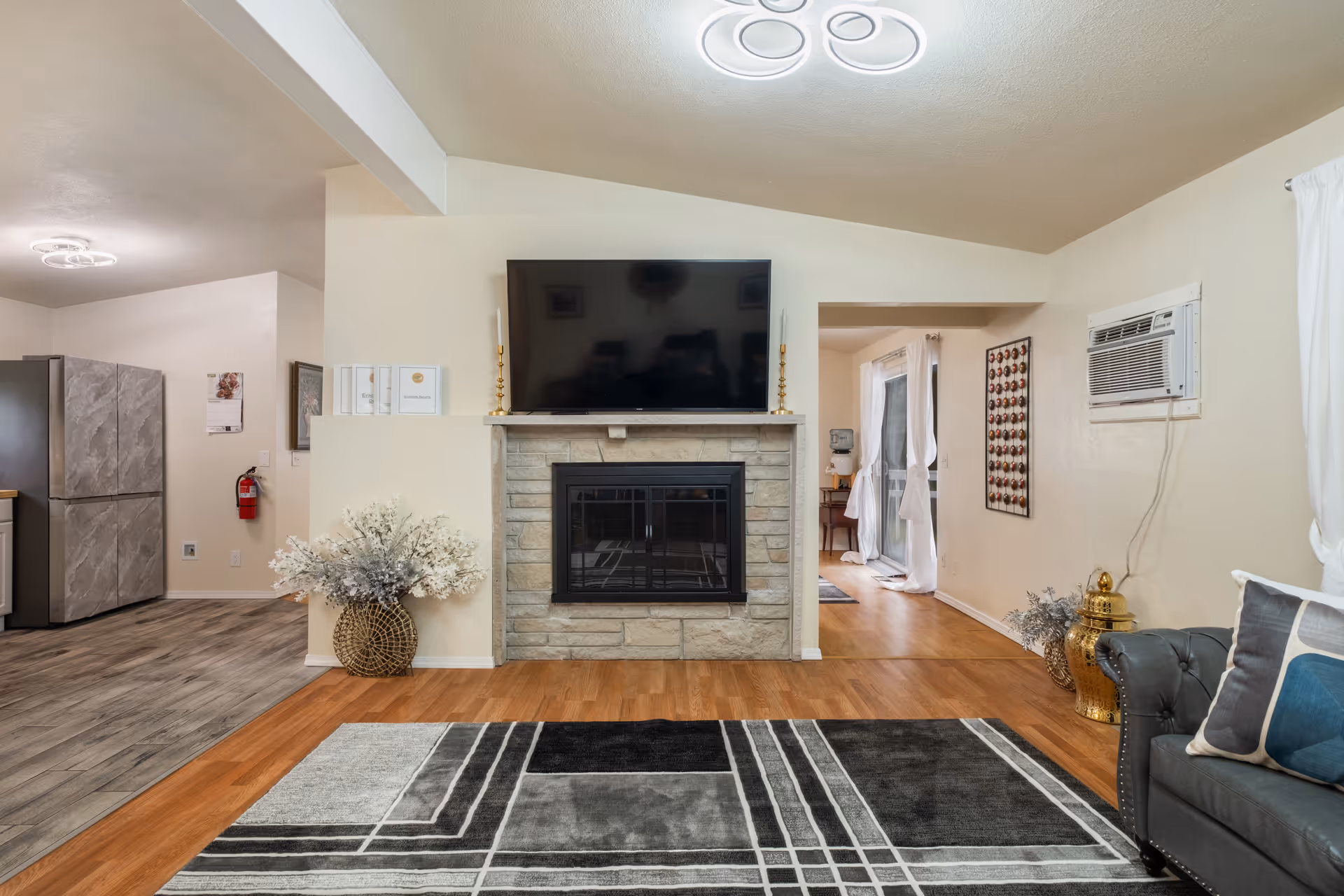 A cozy living room with a stone fireplace and a flat-screen TV mounted above it. The room features a patterned area rug on a wooden floor, a gray couch with decorative pillows on the right, and a large vase with white flowers on the left. The adjacent kitchen area with a refrigerator is visible to the left, and a hallway with natural light coming through sheer curtains is seen to the right. The walls are painted light beige, and there is an air conditioning unit mounted on the right wall.