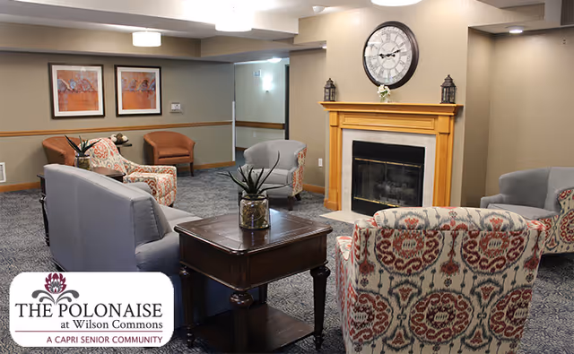 Comfortable senior-living common room with armchairs arranged around a coffee table facing a fireplace topped by a clock.