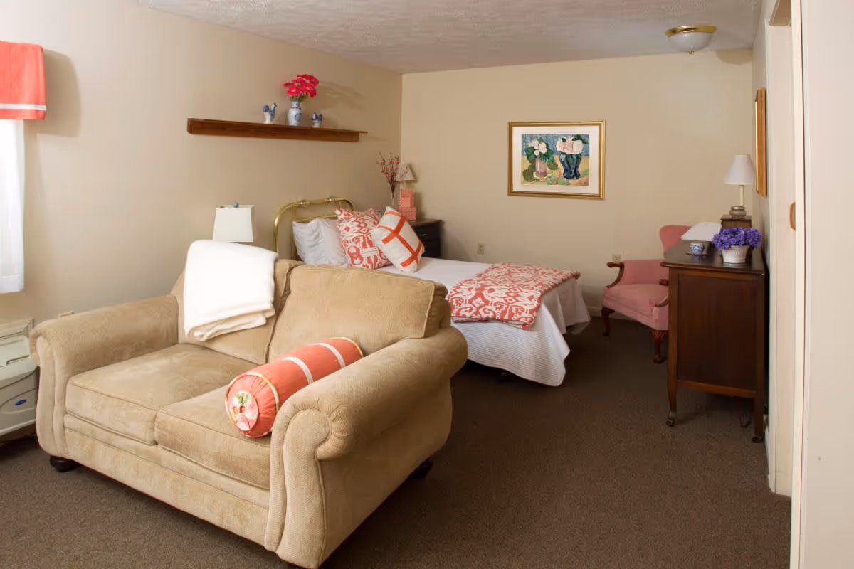 A cozy bedroom in a senior living facility featuring a beige loveseat with a white blanket and a coral bolster pillow in the foreground. Behind it is a bed with white and coral bedding and decorative pillows. The room has beige walls, a framed floral painting, a wooden dresser with a lamp and purple flowers, and a pink upholstered chair. A wooden shelf with decorative items is mounted on the wall above the bed.