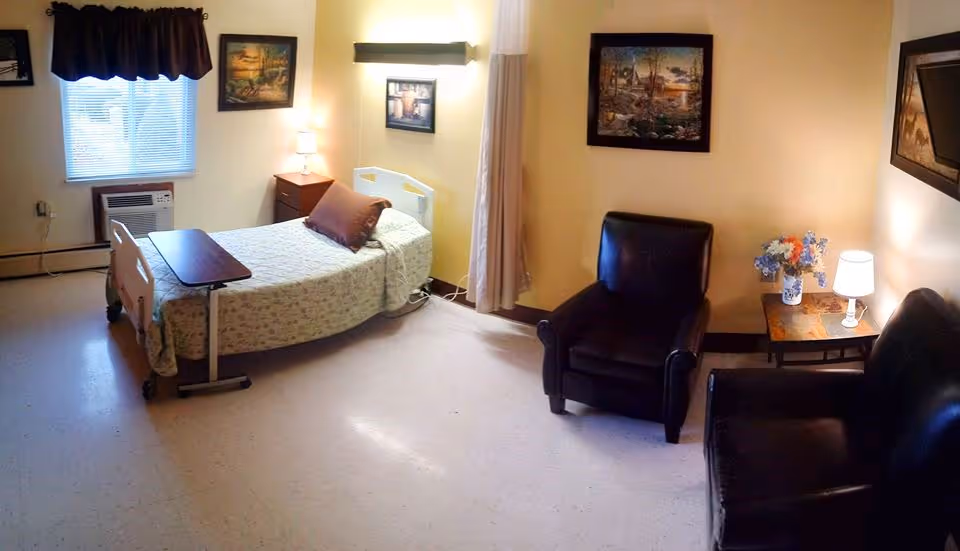A cozy room with a single hospital-style bed covered with a floral bedspread and a brown pillow. Next to the bed is a small wooden nightstand with a lamp. There is a window with blinds and a dark valance. The room also features two black leather chairs, a small wooden side table with a lamp and a vase of flowers, and framed artwork on the walls. The floor is light-colored and the walls are painted a soft yellow.