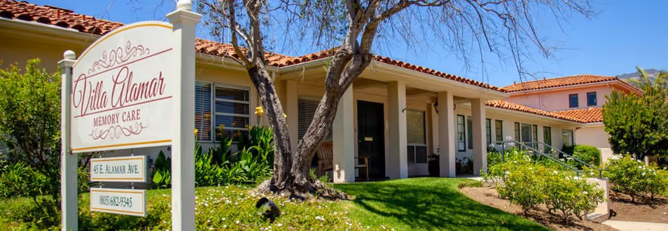 Exterior view of Villa Alamar Memory Care facility showing a single-story building with a red tile roof, a green lawn, trees, and a white sign with the facility name, address, and phone number.