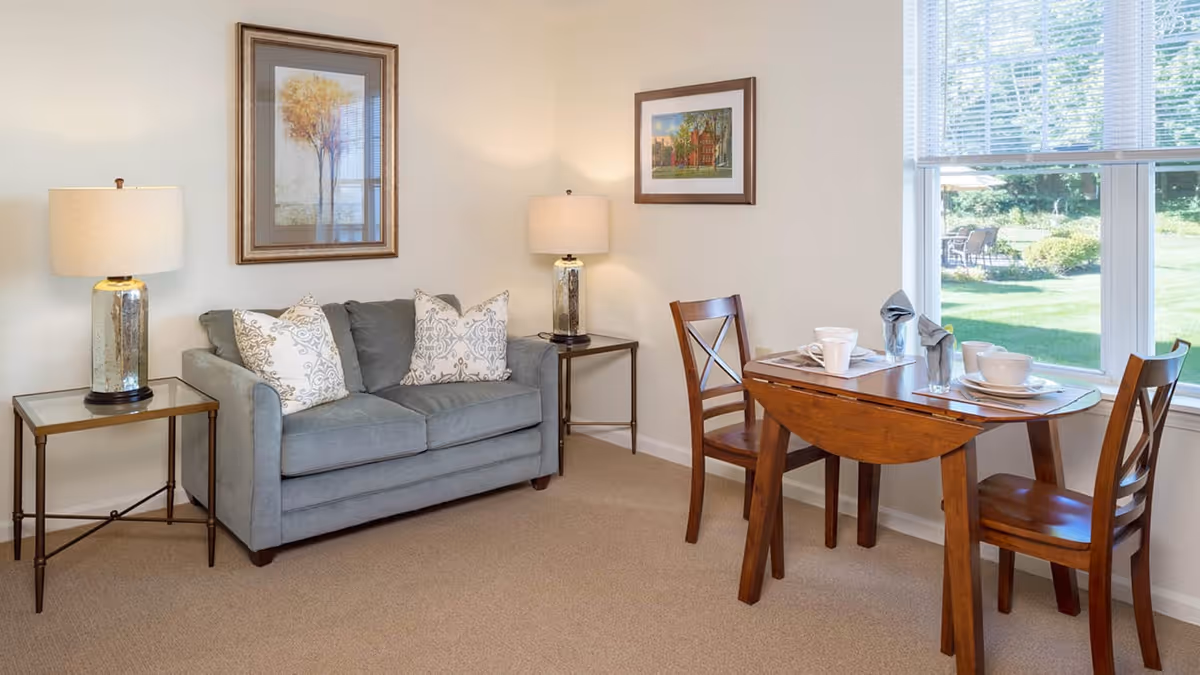 A cozy living area with a gray loveseat adorned with patterned pillows, two side tables each holding a lamp, a small wooden dining table set for two with dishes and napkins, and a large window showing a green outdoor space.