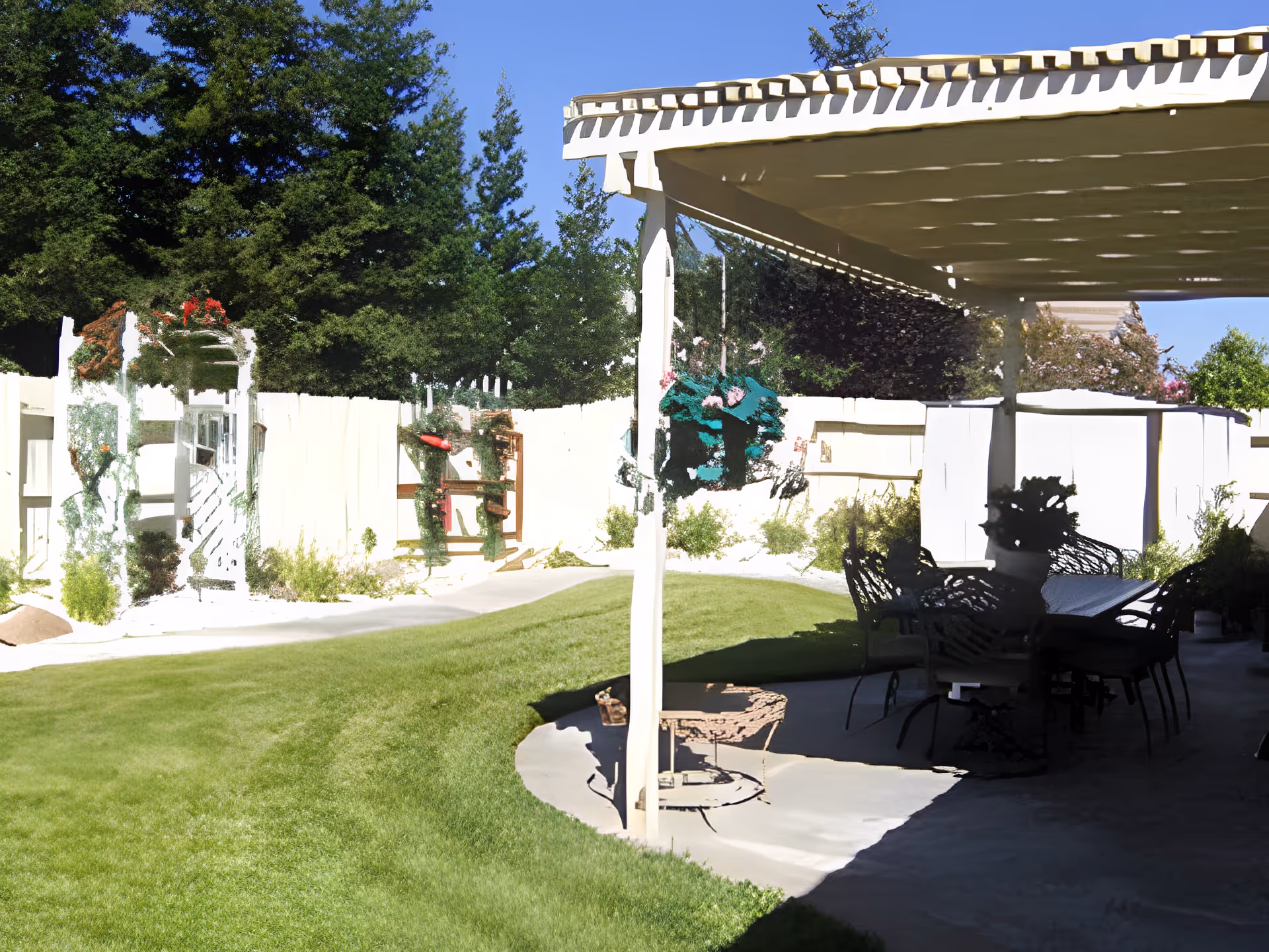 A sunny outdoor garden area with a well-maintained green lawn, surrounded by trees and bushes. There is a covered patio with outdoor furniture including chairs and a table, providing shaded seating. The area is enclosed by a white fence with some decorative plants and flowers.