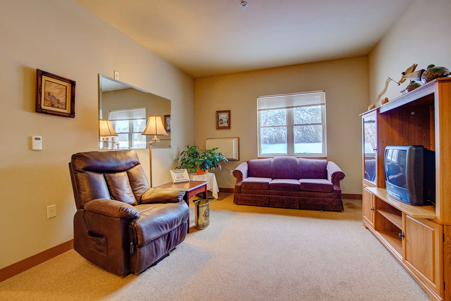 A cozy living room with a brown leather recliner chair, a wooden side table with a lamp and a framed photo, a green potted plant, a purple upholstered sofa under a window with blinds, and a wooden entertainment center holding a small TV and decorative items.