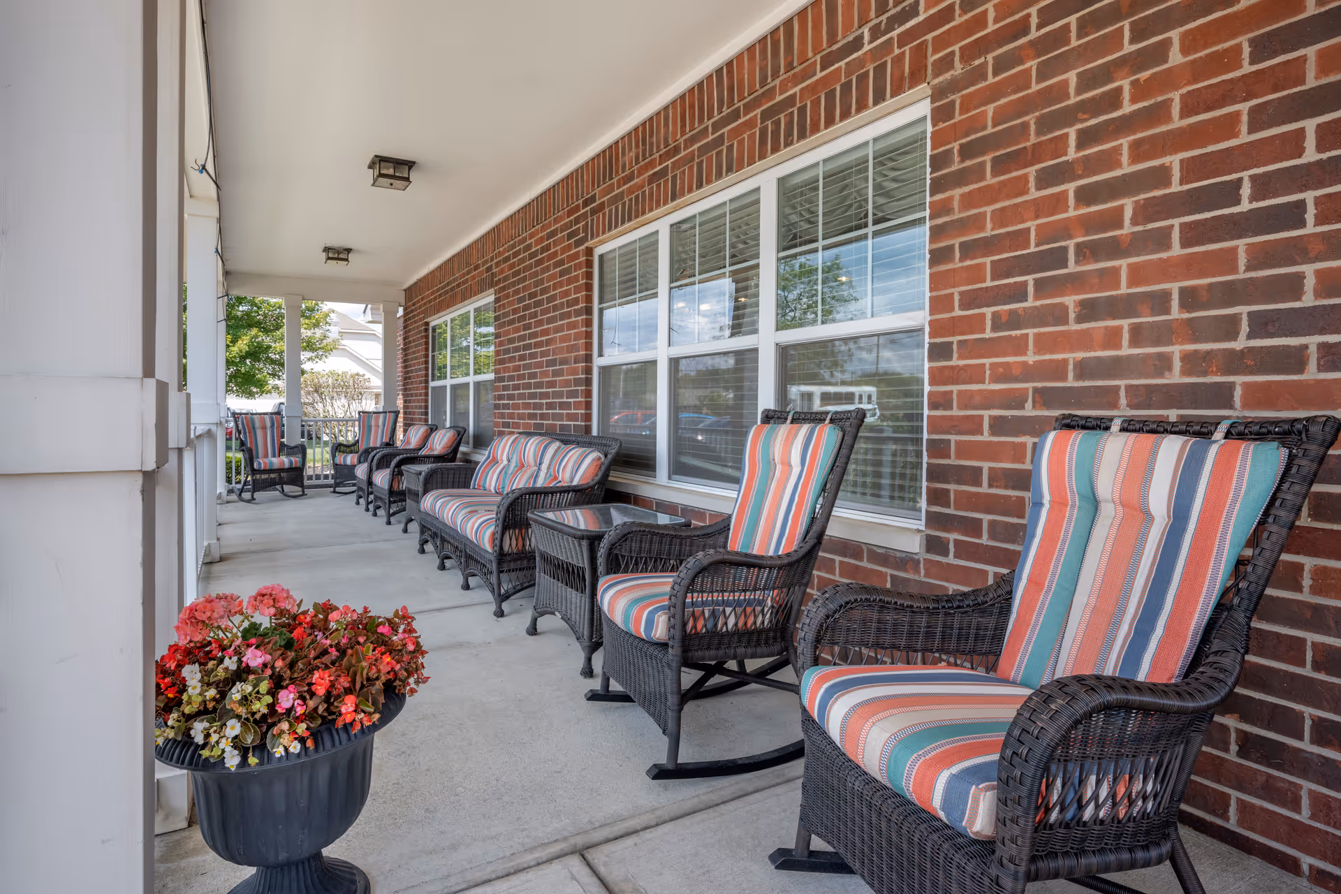 A covered outdoor porch area with a row of wicker rocking chairs and a loveseat, all with striped cushions in shades of red, blue, and white. There is a small glass-top side table between the chairs. A large planter with red and pink flowers is positioned near the entrance. The porch has a brick wall with large windows and white pillars supporting the roof.