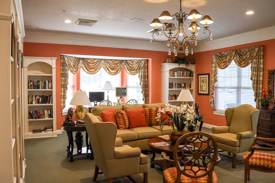 A cozy living room with beige armchairs and a sofa adorned with red and plaid cushions. The room features a wooden coffee table with a flower arrangement, two side tables with lamps, built-in bookshelves filled with books, and windows with plaid valance curtains. A decorative chandelier hangs from the ceiling.
