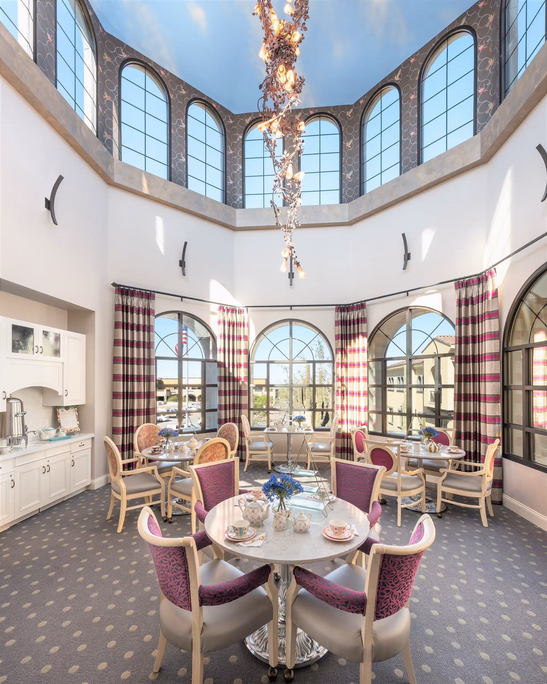 A bright and elegant dining room with large arched windows letting in natural light. The room features round tables with chairs upholstered in purple and beige fabric. Each table is set with teacups, saucers, and a small flower arrangement. The ceiling is high with additional arched windows near the top and a decorative chandelier hanging down. The walls are white with patterned curtains in shades of purple and beige. There is a white cabinet with a coffee urn and cups on the left side of the room.