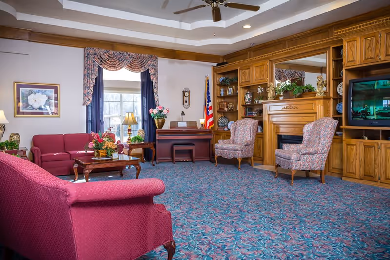 A cozy senior living facility common room with floral patterned carpet, two upholstered armchairs, a red sofa, a wooden coffee table with a flower arrangement, a piano, an American flag, and a large wooden built-in cabinet with a fireplace and a television.