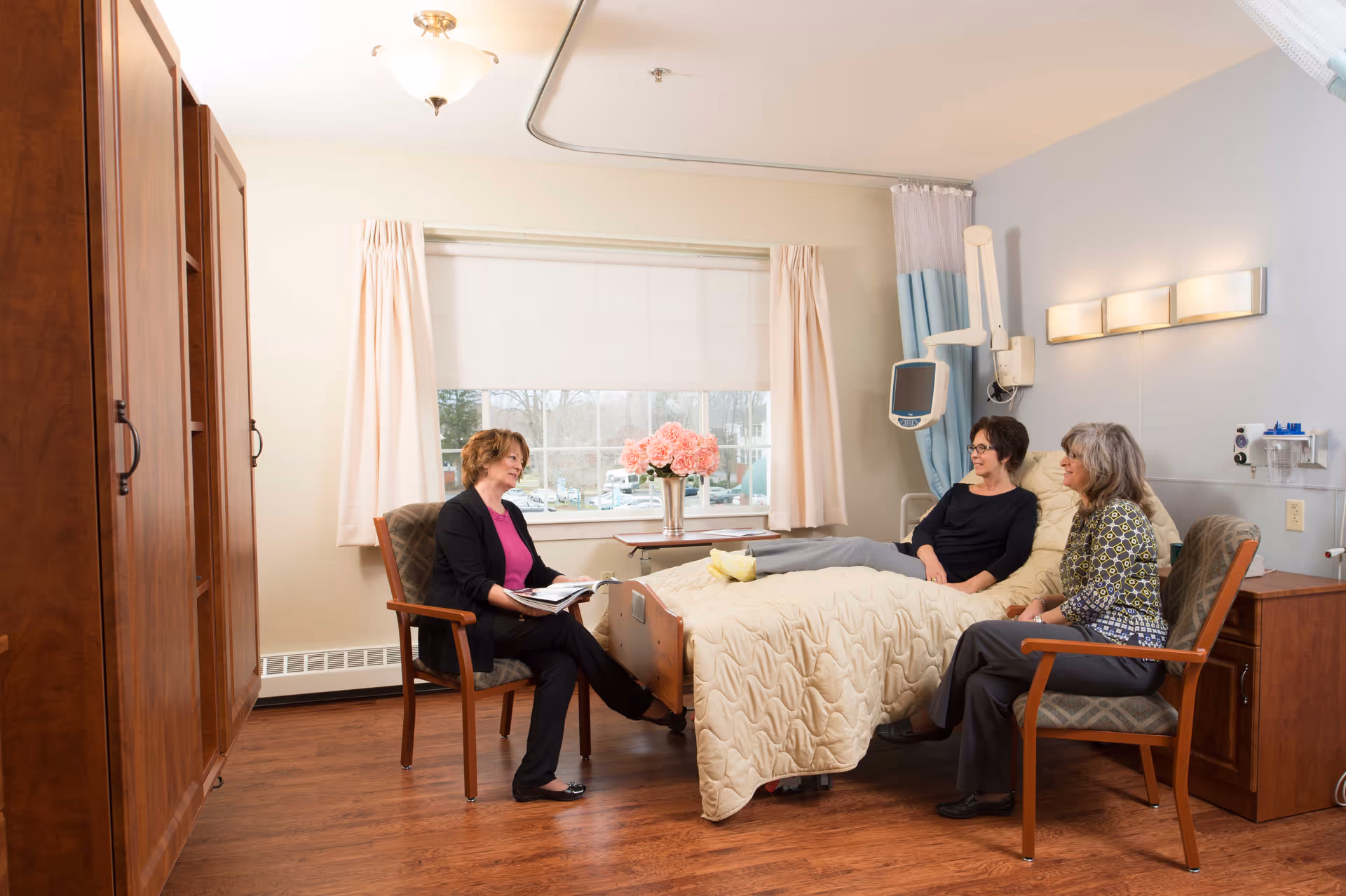 A well-lit patient room in a senior living facility with three women. One woman is sitting on a chair holding a book, while another woman is sitting on a hospital bed with a beige quilted blanket, and the third woman is seated on another chair beside the bed. The room has wooden furniture, a large window with curtains, a vase of pink flowers on a table, and medical equipment mounted on the wall.