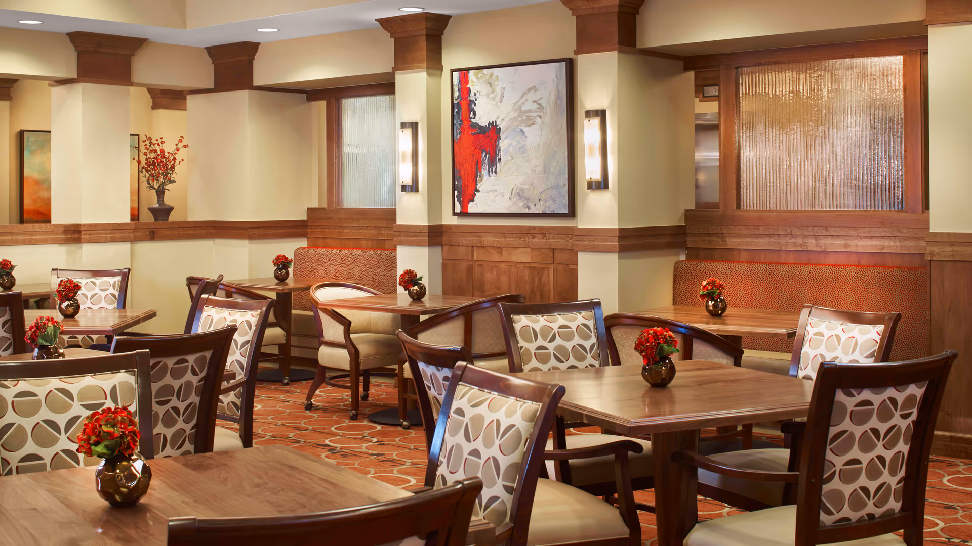 A warm and inviting dining room with wooden tables and chairs featuring patterned upholstery. Small vases with red flowers are placed on each table. The room has beige walls with wooden paneling and decorative columns, a patterned carpet, and framed abstract artwork on the walls.