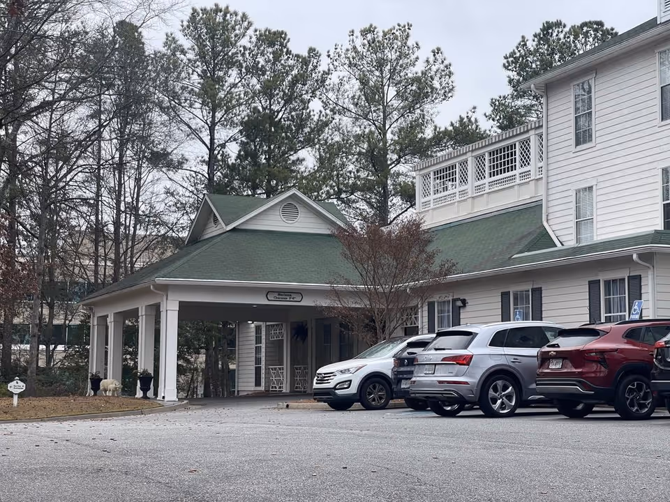 Front entrance porte-cochere of a white multi-story senior living building with parked cars and pine trees.