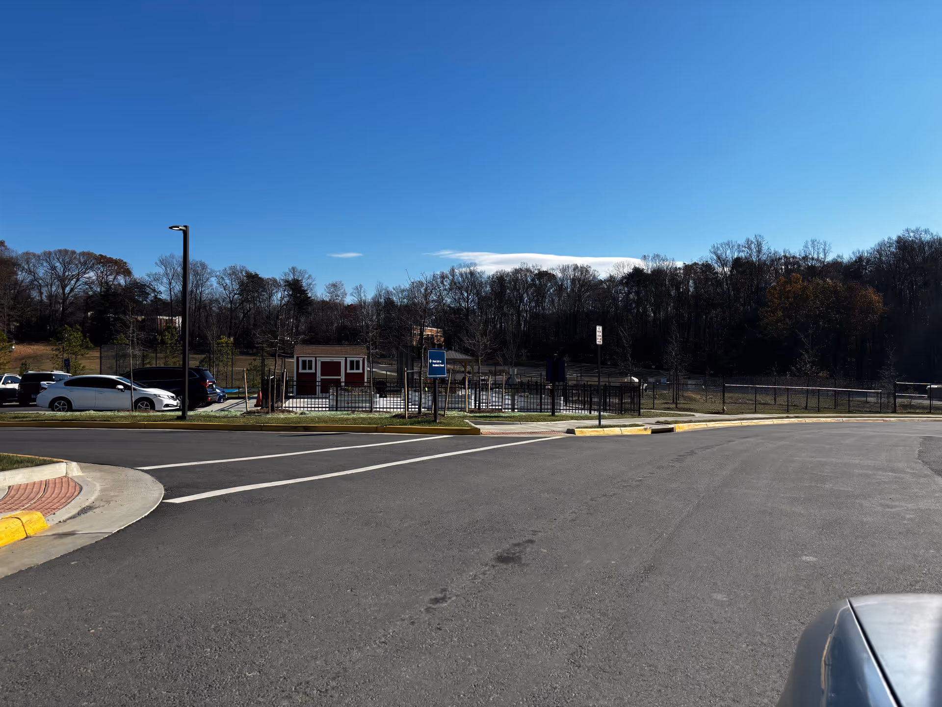 A paved road with a parking area on the left side, several parked cars, a fenced area with a small red and white shed, and trees in the background under a clear blue sky.