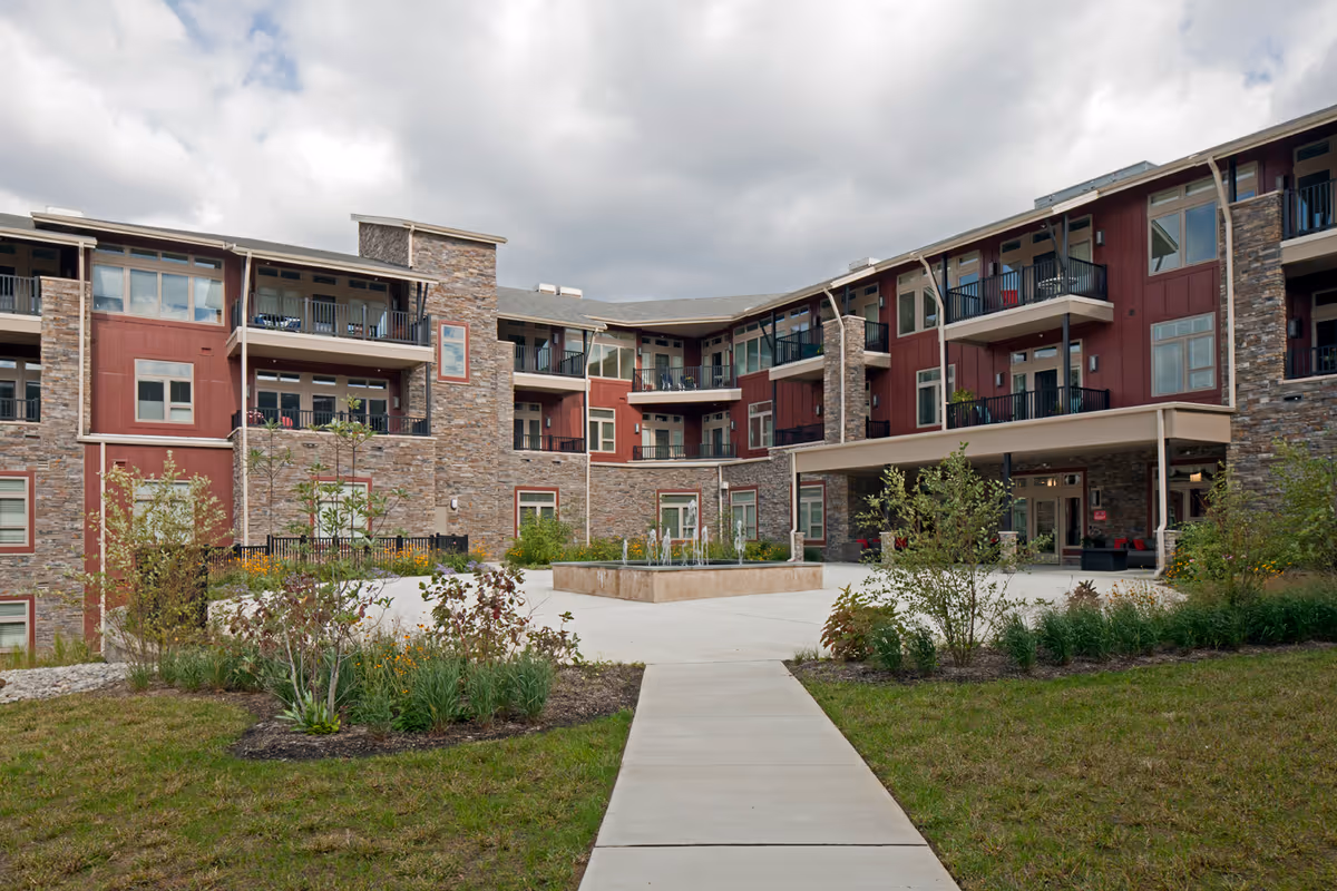 Exterior courtyard view of a three-story senior living building with balconies, stone and red siding and a central walkway.