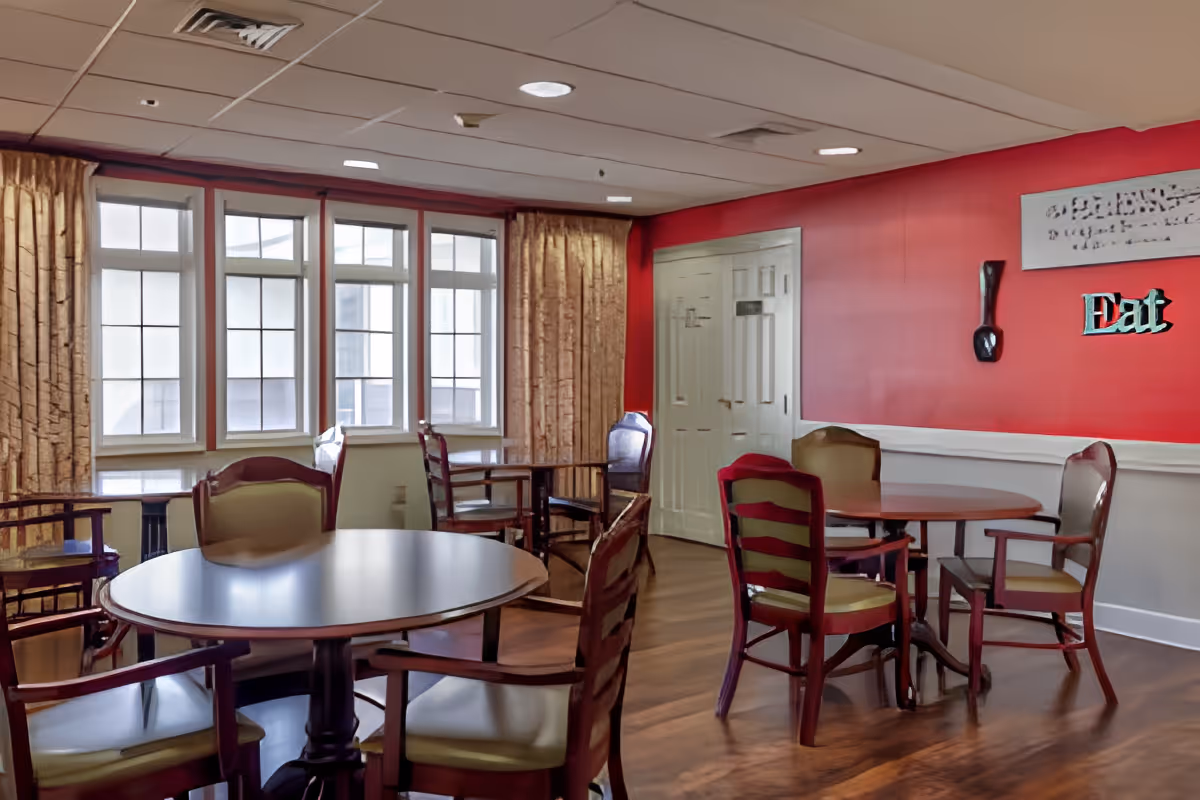 Communal dining room with round tables and wooden chairs, large windows, and a red accent wall with an "Eat" sign.