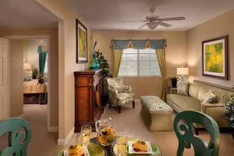 Cozy senior living room with sofa, chairs, ottoman, dining table, window blinds, ceiling fan, and artwork.