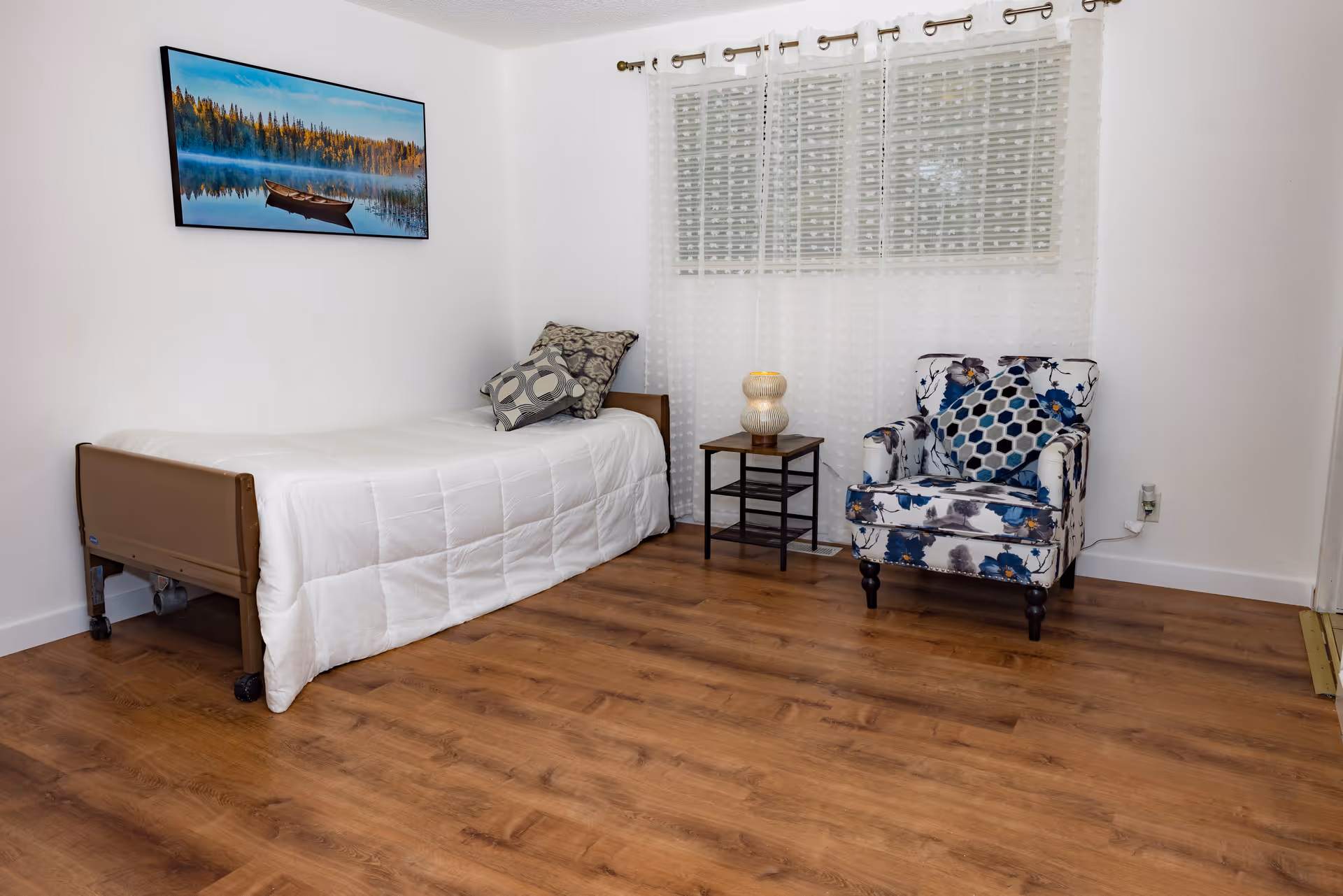 Small furnished bedroom with a single bed, patterned armchair, a side table with lamp, wall art and a window with sheer curtains.