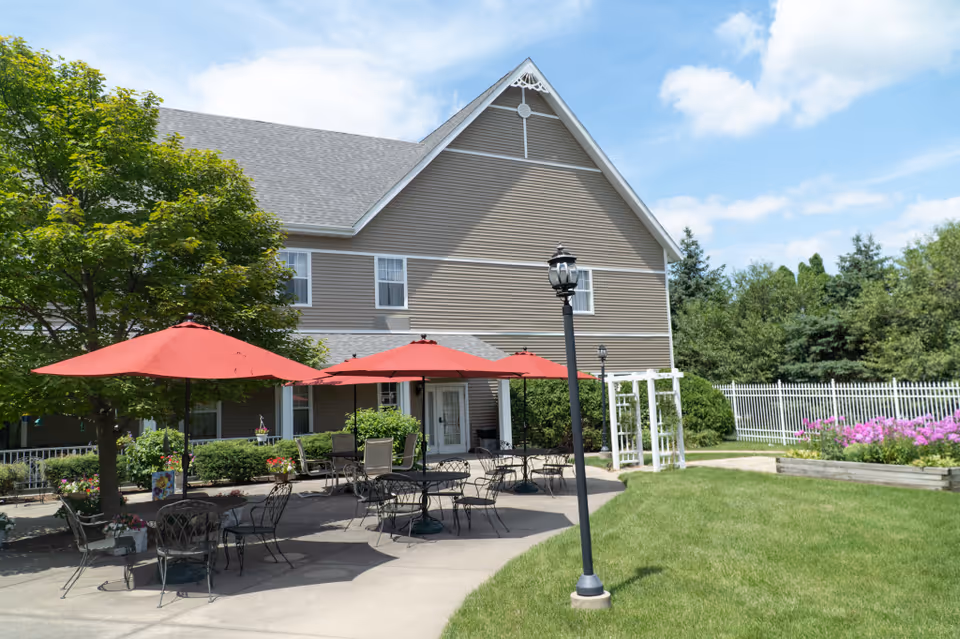 Outdoor patio area at a senior living facility with several black metal tables and chairs under red umbrellas. The patio is adjacent to a beige building with white trim, surrounded by green trees, bushes, and a white fence. There are also flower beds with pink flowers and a clear blue sky with some clouds above.