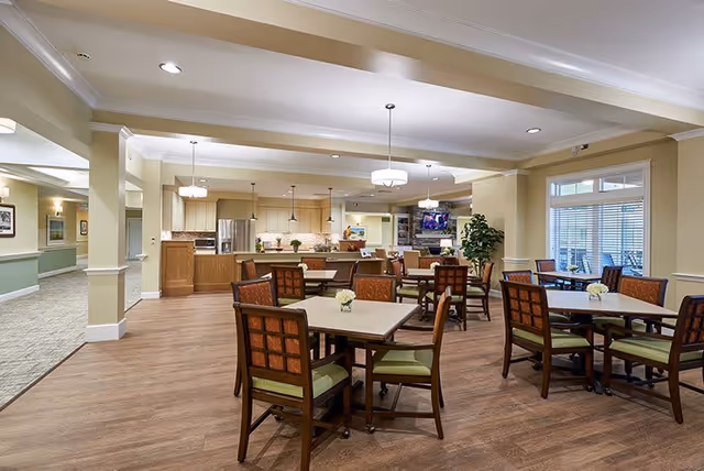 A spacious and well-lit dining area in a senior living facility featuring multiple square tables with four chairs each. The chairs have wooden frames with green cushions and patterned backs. The room has light-colored walls, wood flooring, and modern pendant lights hanging from the ceiling. In the background, there is a kitchen area with stainless steel appliances and a countertop, as well as a seating area with a television mounted above a stone fireplace. Large windows with blinds allow natural light to enter the space.