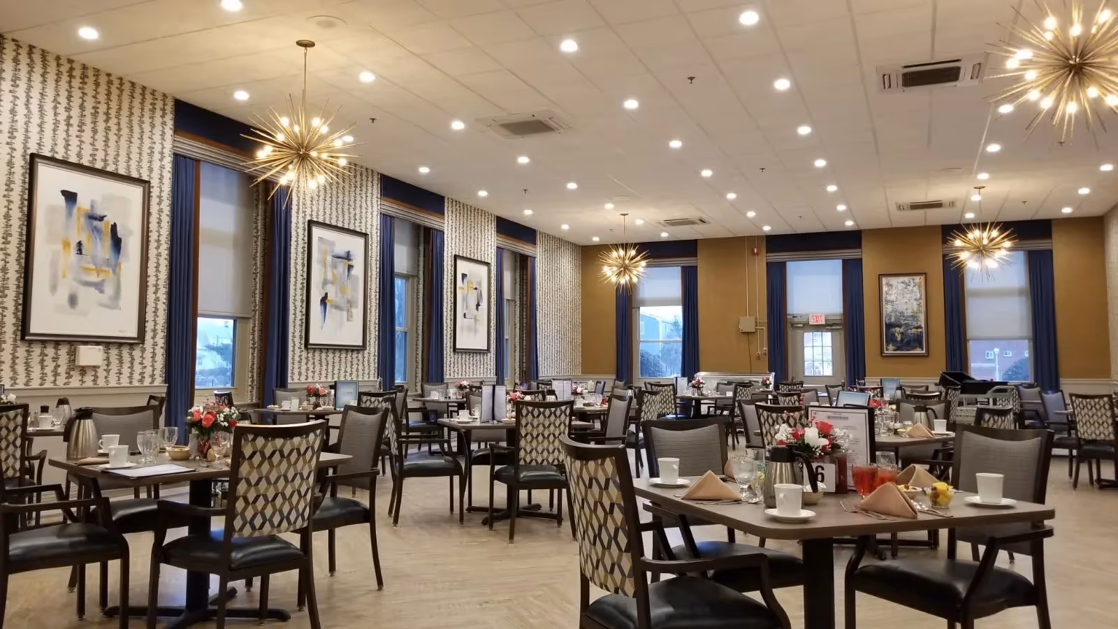 A spacious and well-lit dining room with multiple tables and chairs arranged neatly. Each table is set with cups, glasses, napkins, and small flower arrangements. The room features large windows with blue curtains, patterned wallpaper, modern starburst chandeliers, and framed abstract artwork on the walls.