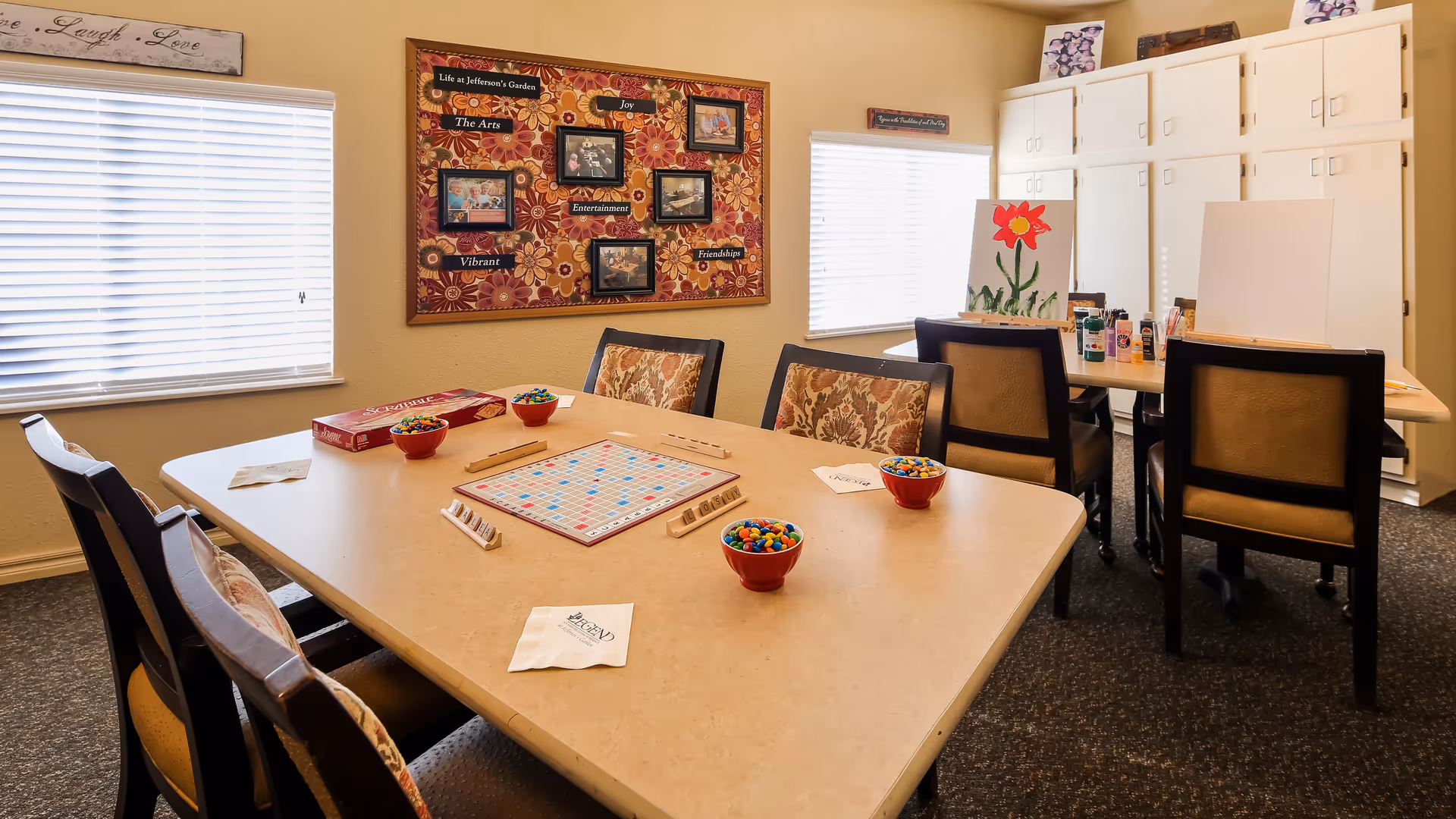 A well-lit activity room with two tables and several chairs. One table is set up for a Scrabble game with tiles, racks, and bowls of candy. The other table has painting supplies, including canvases, paint bottles, and a painting of a red flower. The walls have windows with blinds, a decorative bulletin board with framed photos and words like 'Joy' and 'Friendship,' and cabinets for storage.