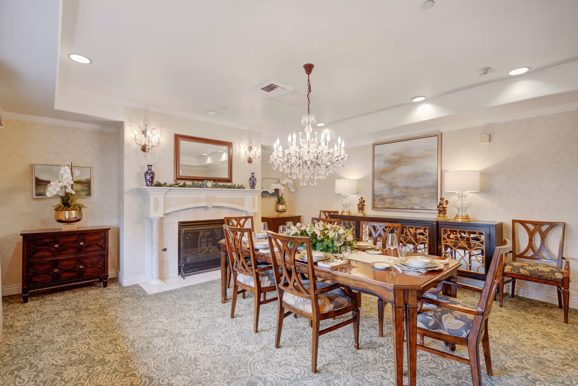 Elegant dining room with a wooden dining table set for six, surrounded by matching wooden chairs with patterned cushions. A large crystal chandelier hangs above the table. The room features a fireplace with a mirror above it, two wall sconces, a sideboard with decorative lamps, and a large framed painting on the wall. The carpet has a floral pattern, and the walls are decorated with light wallpaper.