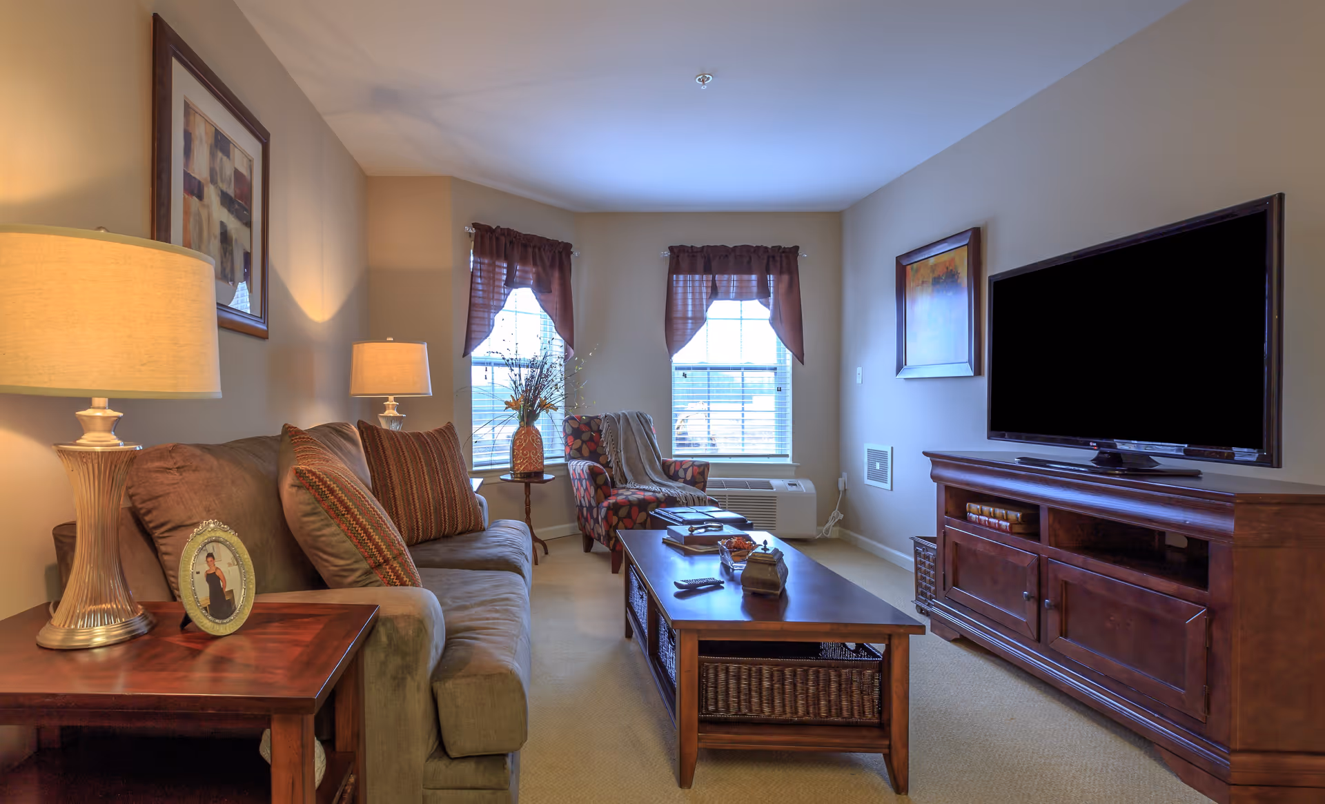 A cozy living room with a brown sofa adorned with striped pillows, a wooden coffee table with decorative items, a patterned armchair near two windows with brown curtains, a wooden TV stand with a flat-screen television, two table lamps providing warm lighting, and framed artwork on the walls.