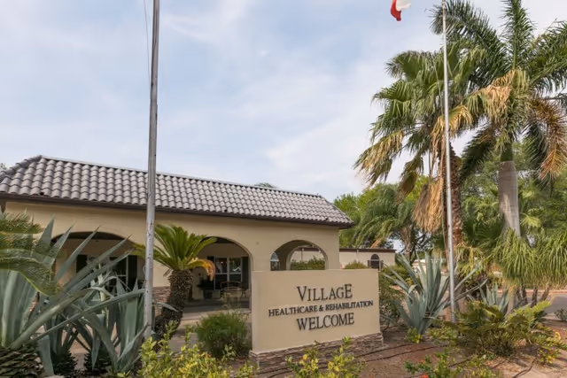 Exterior view of Village Healthcare and Rehabilitation facility with a tiled roof building, palm trees, and a sign that reads 'Village Healthcare & Rehabilitation Welcome' in front of the entrance.