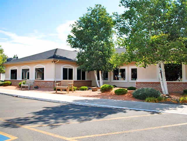 Front exterior of a single-story assisted living building with benches, landscaped trees, and a parking lot.