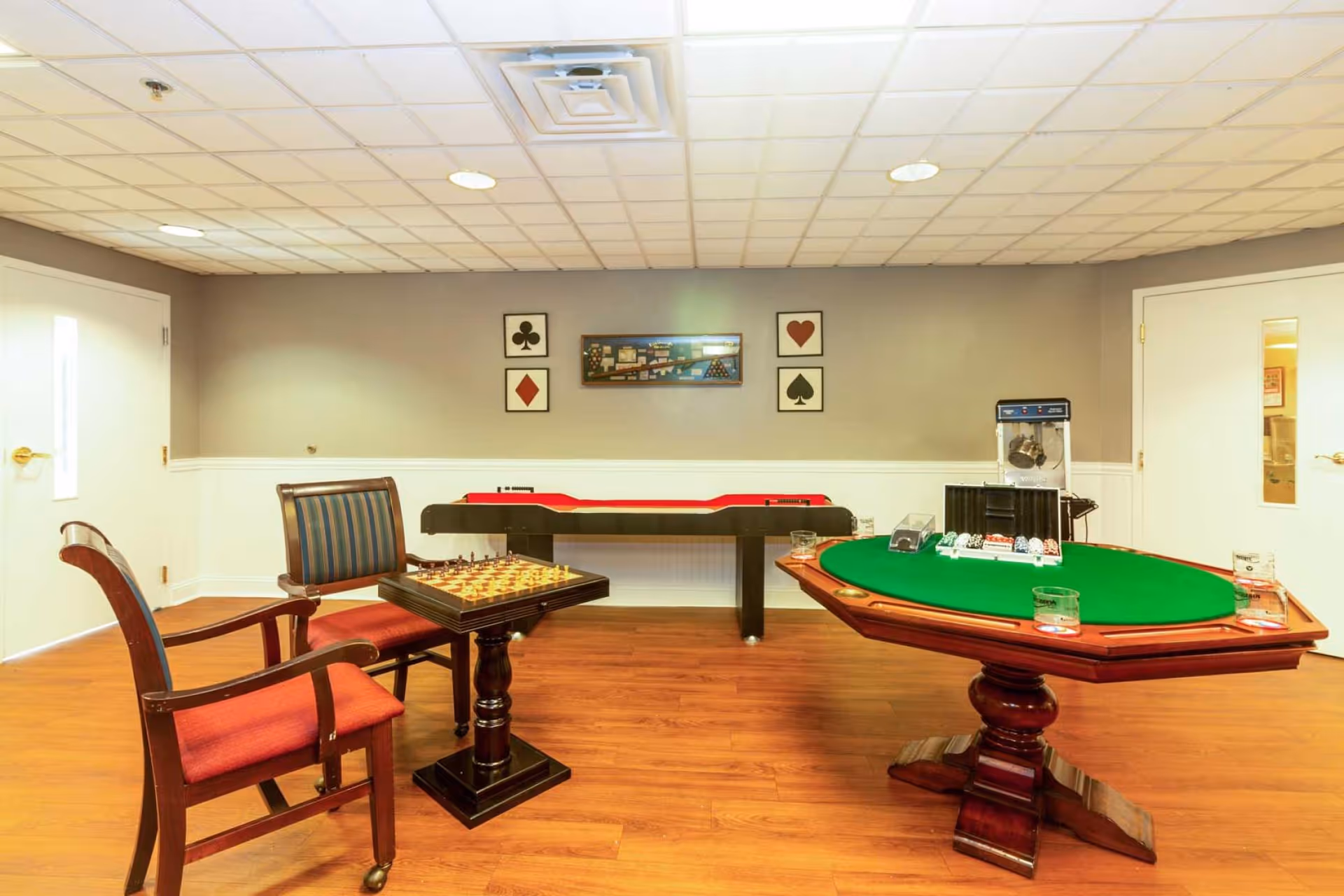 A game room with a wooden floor and a white ceiling. The room contains a small chess table with two wooden chairs, a shuffleboard table against the wall, and a poker table with green felt and poker chips. The walls are painted gray with white wainscoting, and there are framed pictures of playing card suits and a pool table on the wall.