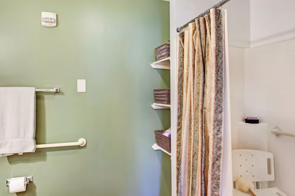 Bathroom with a green wall, a white towel hanging on a grab bar, a toilet paper holder, built-in shelves with brown baskets, and a shower area with a striped shower curtain and a white shower chair.