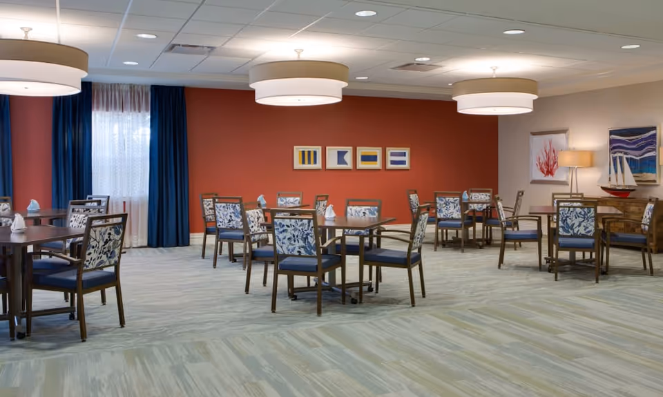 A spacious dining room with multiple wooden tables and chairs featuring blue and white floral upholstery. The room has a red accent wall with framed nautical-themed artwork, blue curtains, and large round ceiling lights. There is also a sideboard with a model sailboat and additional artwork on the walls.